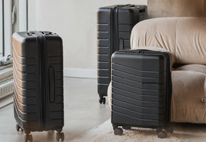 Three black suitcases of different sizes on a carpeted floor with a beige sofa in the background.
